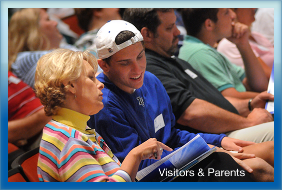 a male student seaated with his mother at an audience of parents and students with words saying Visitors and Parents