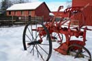 red farm plow sitting in the snow by a red barn.