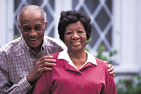 An older man and woman standing in front of a house.  Both are smiling into the camera.  The man is standing behind the woman with his hands on her shoulders.