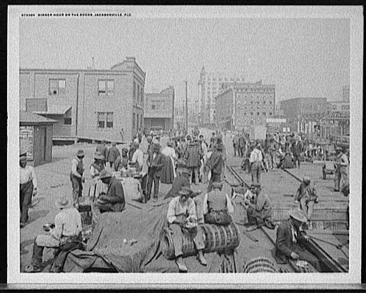 Black and white image of dozens of male workers sitting on barrels or on the ground eating dinner at a dock in Jacksonville, Florida.     