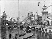 A walkway over water is at the center of the photograph.  A giant roller coaster and buildings built in the style of castles are in the background.