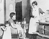 A young woman is seated with a baby on her lap in the center of the photo.  On the right are two young children.  One child is standing.  The other is seated in a crib.  A woman in a long white apron stands by the stove on the left side of the photo.  She is pulling a bottle out of a pan on the stove.  