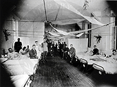 Black and white photo of rows of beds.  Patients sit on the beds. 