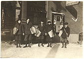 A black and white image of 5 boys, each about 10 years of age, standing outside of a bank holding stacks of newspapers to sell, c1904 