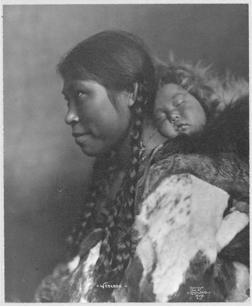 An Eskimo mother is shown in profile with her child sleeping on her back.