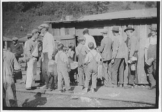 Black and white image of a group of men and boys, some carrying buckets, waiting outside of a building for rations to be distributed.    