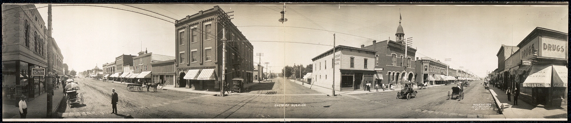 A panoramic view of an intersection in Sheridan, Wyoming.  Stores line the streets and automobiles along with a few horse and buggies are on the road.  