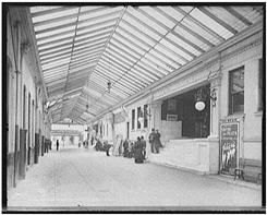 Men and women sitting and standing along one side of the enclosed arcade walkway.  Theatre advertisement and entrance to arcade visible in the foreground.