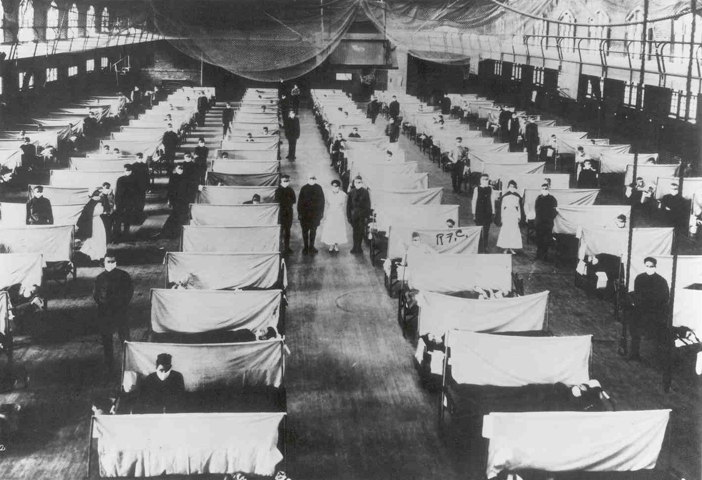 A row of tented beds in a gymnasium.  Doctors and nurses stand by the beds.   