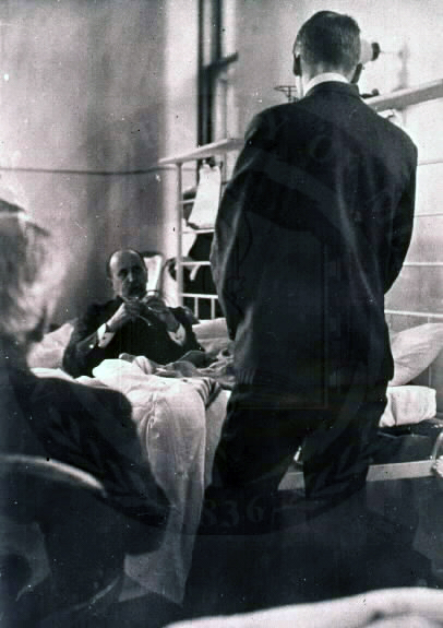 A physician stands with his back to the viewer.  The physician stands next to a bed with a patient.  Another physician is seated on the opposite side of the bed.