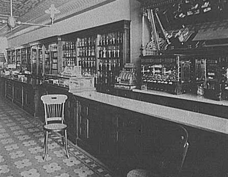 A black and white image of a c1910 drugstore interior. The counter, two chairs, and shelves stocked with medical supplies are visible. 