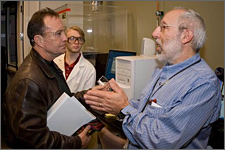 Photo of two men facing each in conversation. On the left, the man has brown hair and is wearing clear safety eyeglasses. He is wearing a brown leather jacket and carrying a bundle of white papers in his hand. He is listening to the man on the right, who is speaking intently and gesturing with his hands clasped together. He has short gray and white hair and a closely trimmed white beard. He is wearing a blue long-sleeved shirt. Behind them stands a row of scientific equipment covered in a clear hard plastic case and linked to a computer.