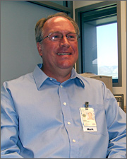 Photo of a man seated at a desk. He is wearing a blue shirt, a badge, and eyeglasses. He is smiling and has short hair.