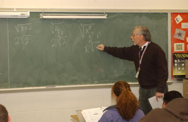 A photo of a male teacher writing math formulas on a chalk board.    