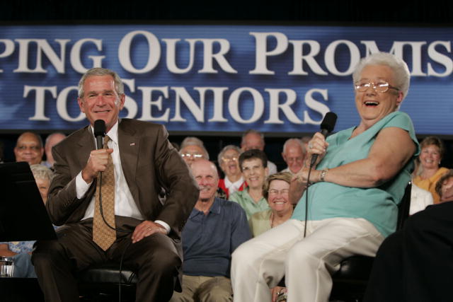 This picture depicts President Bush sitting onstage on a stool with a Medicare beneficiary discussing the Medicare program. The beneficiary is an 82–year-old retired Scottsdale grandmother who was on hand to lend support to the President’s Conversation on Medicare. The picture’s background shows a portion of a blue banner, which reads something like: "Keeping Our Promise to Our Seniors."