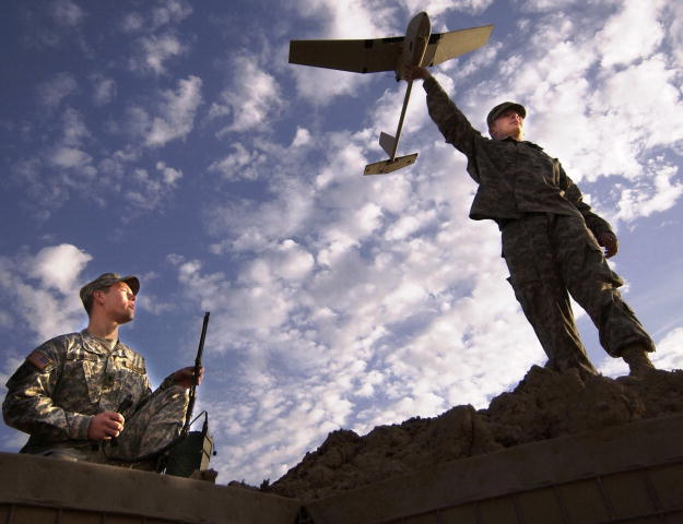 A photo of two male soldiers.  One is sitting on the ground while the other is standing and holding the aerial vehicle above his head.  