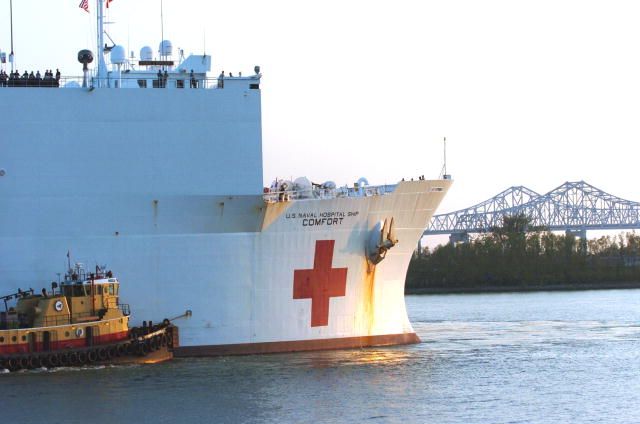 This photo shows the Hospital ship USNS Comfort pulling into port, with a bridge in the background and a tug boat next to the ship.  