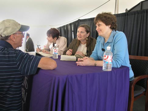Three women on a panel signing books while a man speaks to them.