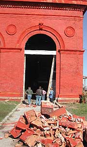 Rubble piled up in front of a large, red, damaged building, with people walking into the building's arched doorway.