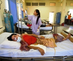 A nurse checks a patient's IV in Cambodia. Male patient is lying in hospital bed with a mask over his face.