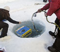 Photo of two people lowering a device into a hole in the ice.