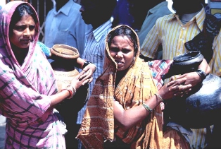 Two Indian women standing in a line holding large water jugs under their arms.