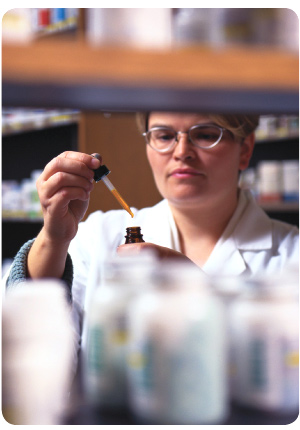 photo of pharmacist in the back area of a pharmacy, adding a liquid to the contents of a small glass bottle with an eye dropper,