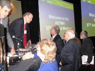 People greeting the lecturers of the symposium with two screens announcing the ASTMH meeting in the background.