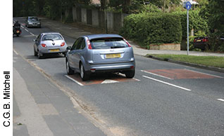 The speed cushions shown here— one directly underneath the closest car and another in the lane to the right of the car—affect automobiles more than larger vehicles. Shortly after this photo was taken, a bus approached in the opposite direction and straddled the speed cushion, leaving its passengers undisturbed.