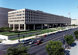 Photo of the U.S. Department of Energy's main building, the Forrestal Building. The large, rectangular office building is lined with windows on all sides and is raised a story above street level by several large pillars.  It is surrounded by a simple lawn and sided by a busy six-lane street.
