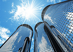 Photo of three office buildings, taken from the base of the buildings.  The tall buildings are covered with shiny glass windows that reflect the clouds and the sky above them.  High above the buildings is the sun.