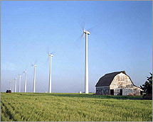 Photo of a string of large, three-bladed wind turbines next to an old barn on a farm.