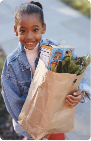 photo of young African-American girl smiling and holding bag of groceries with cereal box and spinach visible at top.