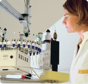 Photo of woman in lab coat looking at numerous glass viles hanging from the arm of a circular piece of lab equipment.