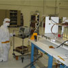 This picture is inside a large 'clean room,' where ATLO (Assembly, Testing, and Launch Operations) is taking place for the Mars Science Laboratory mission. In the foreground on the right are electronic components and to the left is a man leaning over the components. He is dressed in all white protective clothing, including a face mask and gloves.