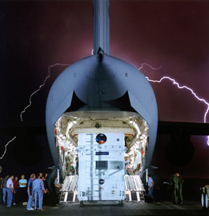 This image shows the tail end of an enormous jumbo jet that is wide open from the back, and 10 people are standing to the sides as a white cargo container with logos of NASA, JPL, and Mars Global Surveyor is rolling out the back of the plane.  The black night sky in the background is electrified with a surprising white lightning streak blazing in a squiggling arc around the C-17.  The lightning turns the sky a deep purple in a haze around the shark-looking tail of the plane.