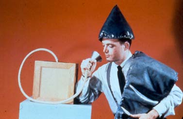 picture of man wearing cone-shaped hat, with pad draped on one shoulder, and holding a funnel attached to tubing going into a box.