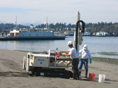 EPA contractors prepare to drive the sample coring tool into the beach to collect a soil sample.  Note the old ferry, the town of Winslow, and the Olympic Mountains in background, highlighting the beautiful setting of the Wyckoff site.