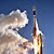 This image features a view from below an Atlas 5 rocket as it lifts off.  A bright blue sky is filled with clouds of smoke from the launch.