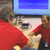 In this image, Graf, a middle-aged Caucasian man with graying, brown hair is on the far left.  He is wearing a bright red shirt and extending his hand to shake that of Flamini. Dr. Flamini, on the right of the image, is a balding Italian man who is smiling broadly.  Behind the men is a screen displaying a blue background with a scratchy white line that represents the successful results of the SHARAD antenna echo test.