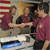 In this image, Graf, a middle-aged Caucasian man with brown hair and a goatee firmly shakes the hand of Naderi, a middle-aged Persian-American man with dark brown hair.  Dr. Charles Elachi, Director of the Jet Propulsion Laboratory (a middle-aged Lebanese-American man) is in the background.  They are all wearing short-sleeved, maroon polo shirts with the mission logo on them.