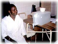 Nurse sitting next to safe water container with a lid and spigot in cholera ward, Guinea Bissau.