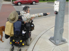 Pedestrian using a wheelchair reaching — unsuccessfully – for a pedestrian pushbutton set up on a curb.