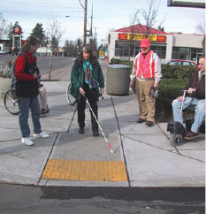 Photo shows orientation and mobility specialist Janet Barlow using a long cane to identify a detectable warning surface retrofitted on an existing curb ramp in a pilot study in Portland, OR. Three other PROWAAC members look on, including Chair Jerry Markesino.