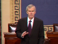 Senator Jeff Bingaman speaking on the floor of the U.S. Senate. He wears a black jacket with dark purple tie, and his hand is poised just above his desk as he emphasizes a point.