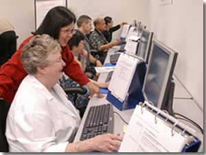 Photo of a classroom instructor leaning over a student assisting with a program on the computer.