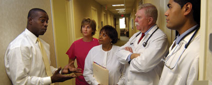 Group photo in the hallway of a medical research building — Hispanic male doctor, Caucasian male doctor, one Black and one Caucasian female nurse, all in white coats or other lab clothing, and Black male statistician in shirt and tie: Statistician speaking to the group with hands extended, describing a research study issue as the others listen.