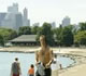 Photo: Man walks on beach with city skyline and trees in background.