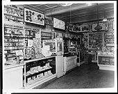 Una foto en blanco y negro del interior de una farmacia. Los mostradores están llenos de artículos y con etiquetas que dicen "Jeringas Fountain" y "Suministros para hospital".