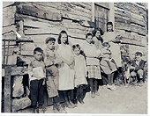 Imagen en blanco y negro de nueve niños entre las edades de 1 a 16 años, recostados sobre el costado de un edificio, algunos de ellos llevan libros de la escuela.